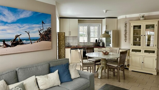 A modern kitchen with white cabinets, a dining table, and chairs. There is a gray sofa with cushions in the living area. - Exclusive apartment with garden and fireplace near the beach (Zingst)