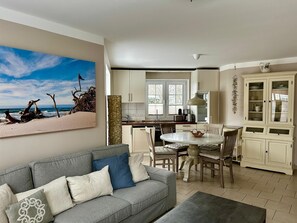 A modern kitchen with white cabinets, a dining table, and chairs. There is a gray sofa with cushions in the living area. - Exclusive apartment with garden and fireplace near the beach (Zingst)