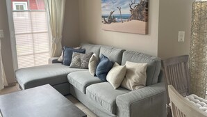 A living room with a grey sectional sofa, a coffee table, and a painting of a beach scene. - Exclusive apartment with garden and fireplace near the beach (Zingst)