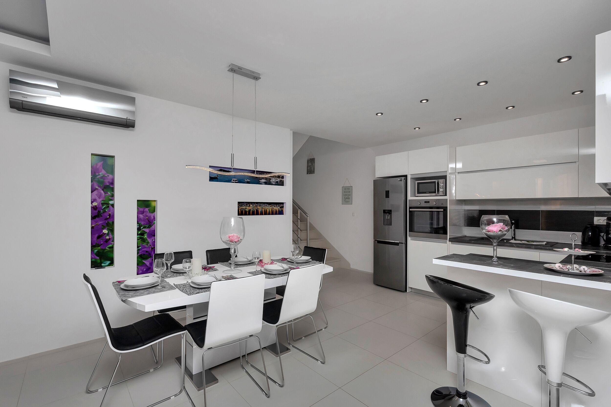 Cozinha moderna com eletrodomésticos de aço inoxidável, área de refeições com mesa branca e cadeiras pretas, e um espaço de bar com banquetas brancas.
