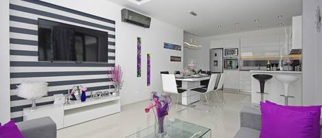 A modern living room with a glass coffee table, a sofa with purple cushions, a dining area with a white table and chairs, and a kitchen with modern appliances.