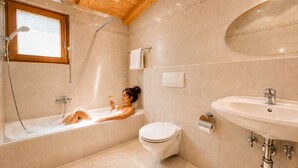 A bathroom with a bathtub, toilet, and sink, wooden ceiling, and beige tiles.
