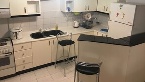 A kitchen with white cabinets, a black countertop, a sink, a stove, two bar stools, and a refrigerator. - Calista Luxury Appt (Kassandra)
