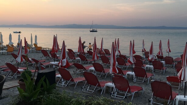 A beach with rows of red and white beach chairs and umbrellas, a boat in the water, and mountains in the background. - Calista Luxury Appt (Kassandra)