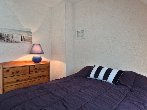 A bedroom with a wooden nightstand, a lamp, a bed with a striped pillow, and a wall with a small picture.