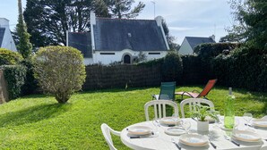 A patio with a round table set for four, surrounded by white chairs, in a lush garden with a hedge and a house in the background.