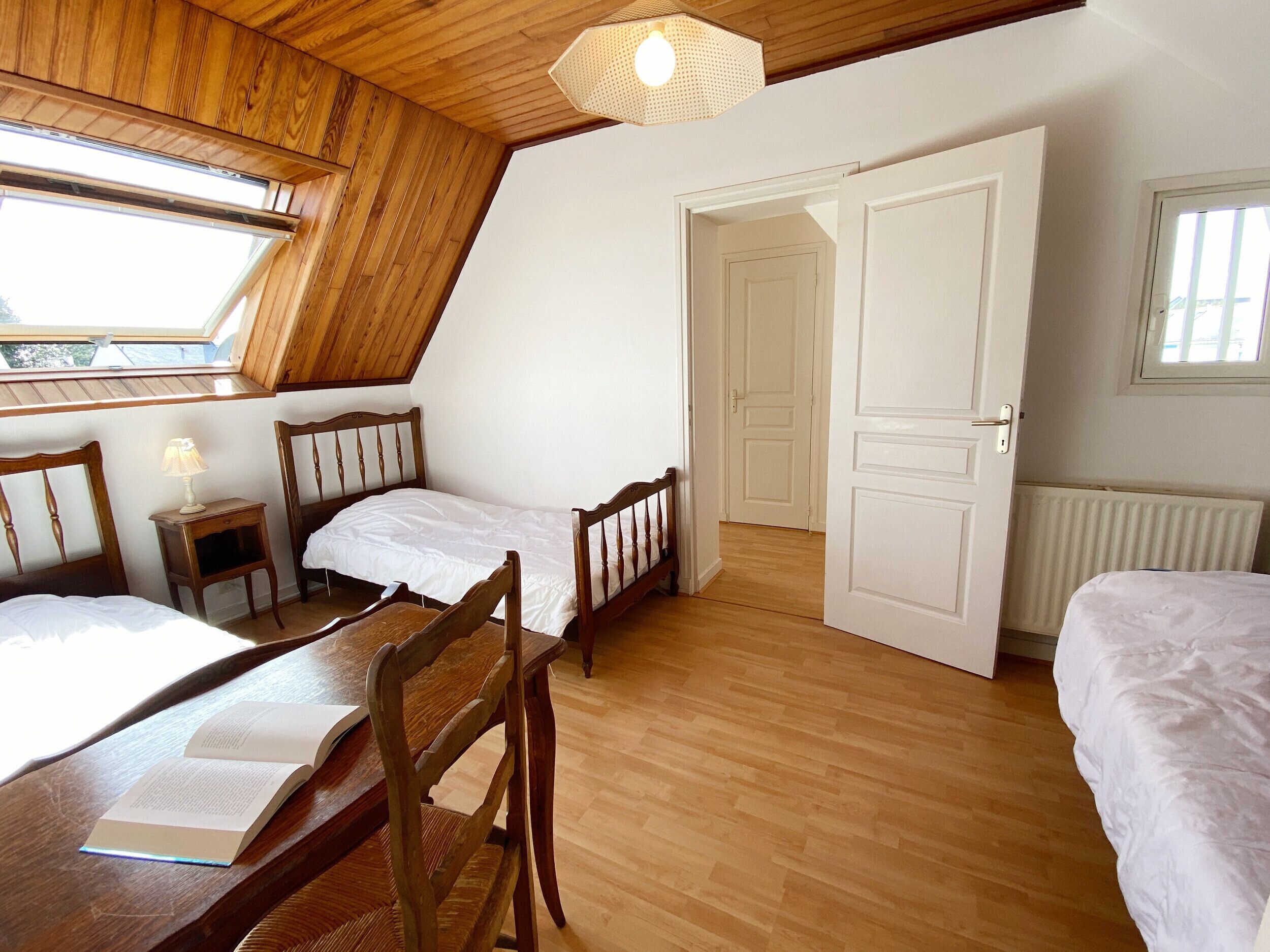A bedroom with two single beds, a wooden desk with a chair, a small bedside table with a lamp, and a large window providing natural light.