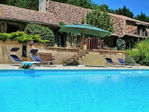 A pool with lounge chairs and an umbrella, surrounded by a building with a tiled roof and steps leading up to it.