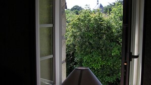 Et værelse med åbent vindue, et bord med lampe og udsigt til grøn natur udenfor.
