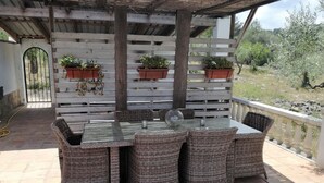 Een terras buiten met een tafel en rieten stoelen, omringd door een houten hek met plantenbakken.