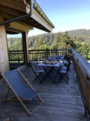 A wooden deck with a table and chairs, a lounge chair, and a view of a forested area.