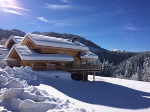 A wooden chalet with a balcony, surrounded by snow-covered trees and a mountain in the background.