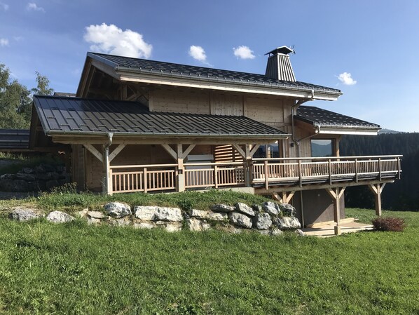 A wooden cabin with a balcony, a chimney, and a stone wall in a grassy area.