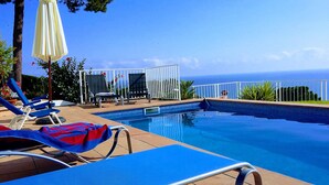 Un espace piscine avec des chaises longues bleues, une clôture blanche et une vue sur l’océan.