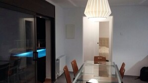 Une chambre d’hôtel moderne avec une table à manger en verre, des chaises en bois, une cabine de douche en verre et un lit avec une tête de lit blanche.