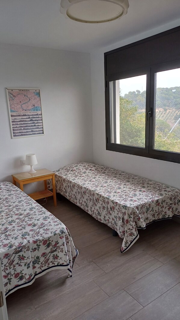 A room with two single beds, a small wooden table with a lamp, a map on the wall, and a large window with a view of trees.