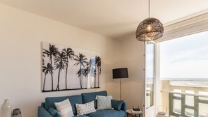 A living room with a blue sofa, a wall-mounted picture of palm trees, a floor lamp, and a view of the sea through a balcony.
