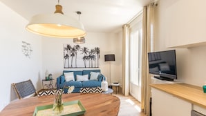 A modern living room with a wooden dining table, a blue sofa with patterned cushions, a television, and a wall-mounted shelf.