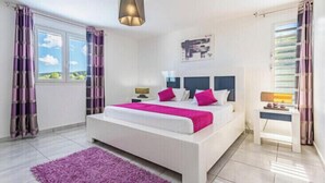 A modern bedroom with a large bed, a bedside table, a nightstand, a window with curtains, and a hanging light fixture. - VILLA BETWEEN SEA AND MOUNTAIN  (Pointe noire)
