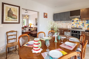 Comedor con mesa de madera dispuesta para cuatro, sillas, un jarrón con flores y una cocina al fondo.