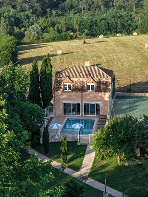 A two-story house with a swimming pool, surrounded by a lush green landscape and trees.