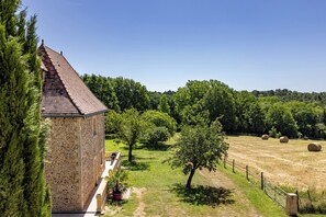A stone building with a tiled roof, surrounded by a grassy field with trees and hay bales.