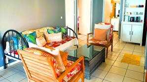 Una sala de estar con sofá, sillón, mesa de centro y una zona de cocina con armarios y electrodomésticos.