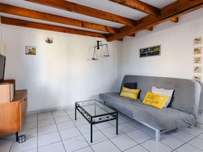 Uma sala de estar com um sofá cinzento, duas almofadas amarelas, uma mesa de centro de vidro, vigas de madeira no teto e um pequeno armário de madeira.