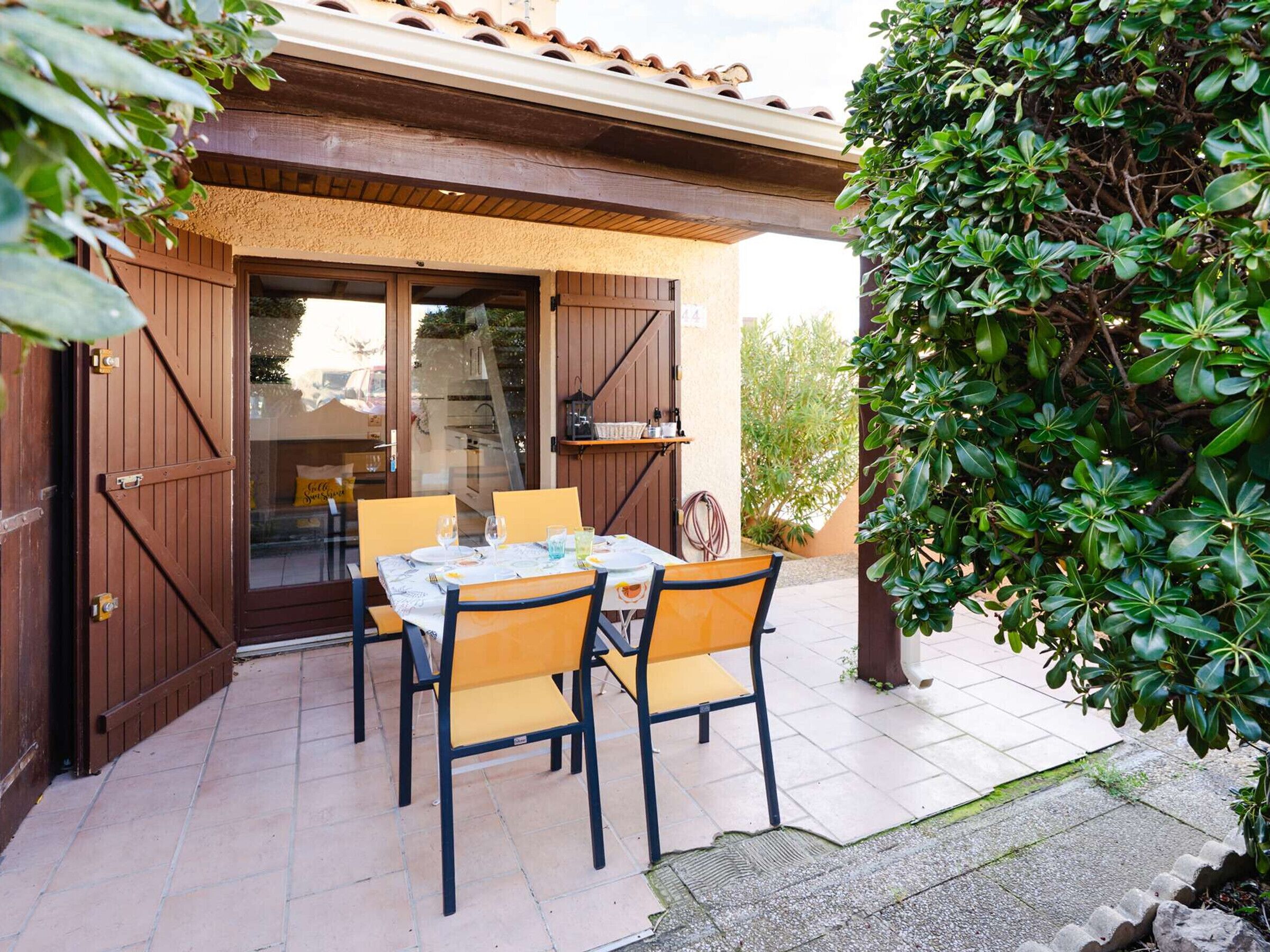 An outdoor patio with a table set for two, surrounded by chairs, and a wooden pergola overhead.