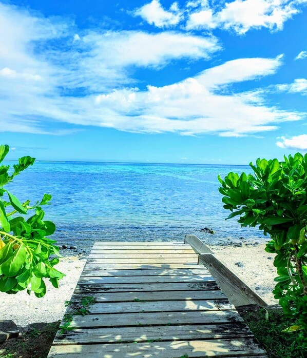 Un sendero de madera que conduce a una playa con agua azul cristalina y un cielo luminoso.
