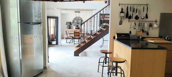 Una cocina con una isla central, taburetes de bar, un colgador para ollas y una escalera que sube a un nivel superior.