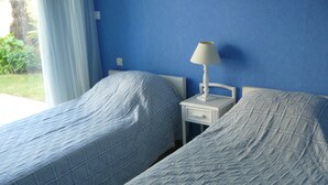 Two single beds with white headboards and nightstands, a white lamp on one nightstand, and a blue wall. - TRIDILLE 1 - Apartment for rent with garden in Carnac - D6 (Carnac)
