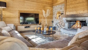 A cozy log cabin interior with a stone fireplace, a television mounted on the wall, and a comfortable seating area with sofas and a coffee table.