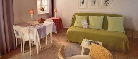 A cozy room with a green sofa, a wicker chair, a small table with a lamp, and a window with curtains.