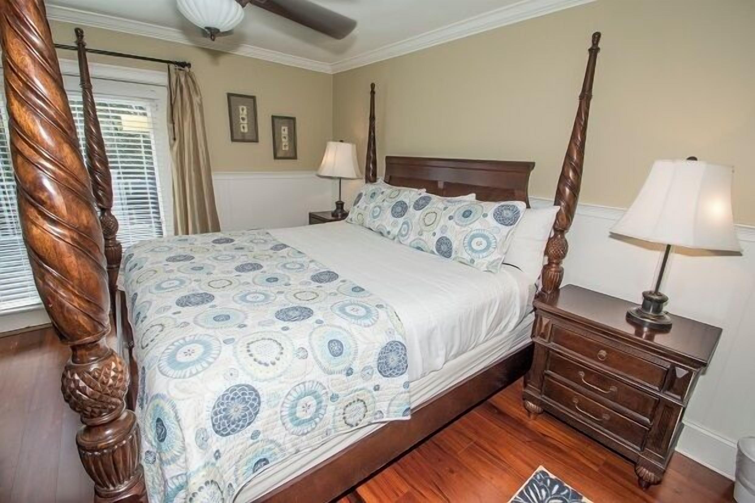 A four-poster bed with a patterned bedspread, a wooden nightstand with a lamp, and a window with blinds.