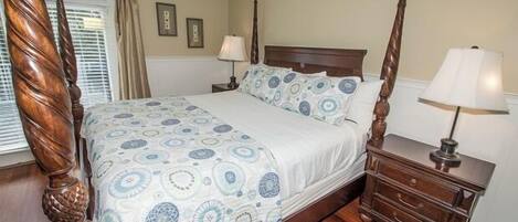 A four-poster bed with a patterned bedspread, a wooden nightstand with a lamp, and a window with blinds.