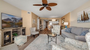 A living room with a TV, seating, a ceiling fan, and a kitchen in the background. - 12th Floor AshWorth, Oceanfront, Wifi, Private Balcony | AshWorth - 1202 (North Myrtle Beach)