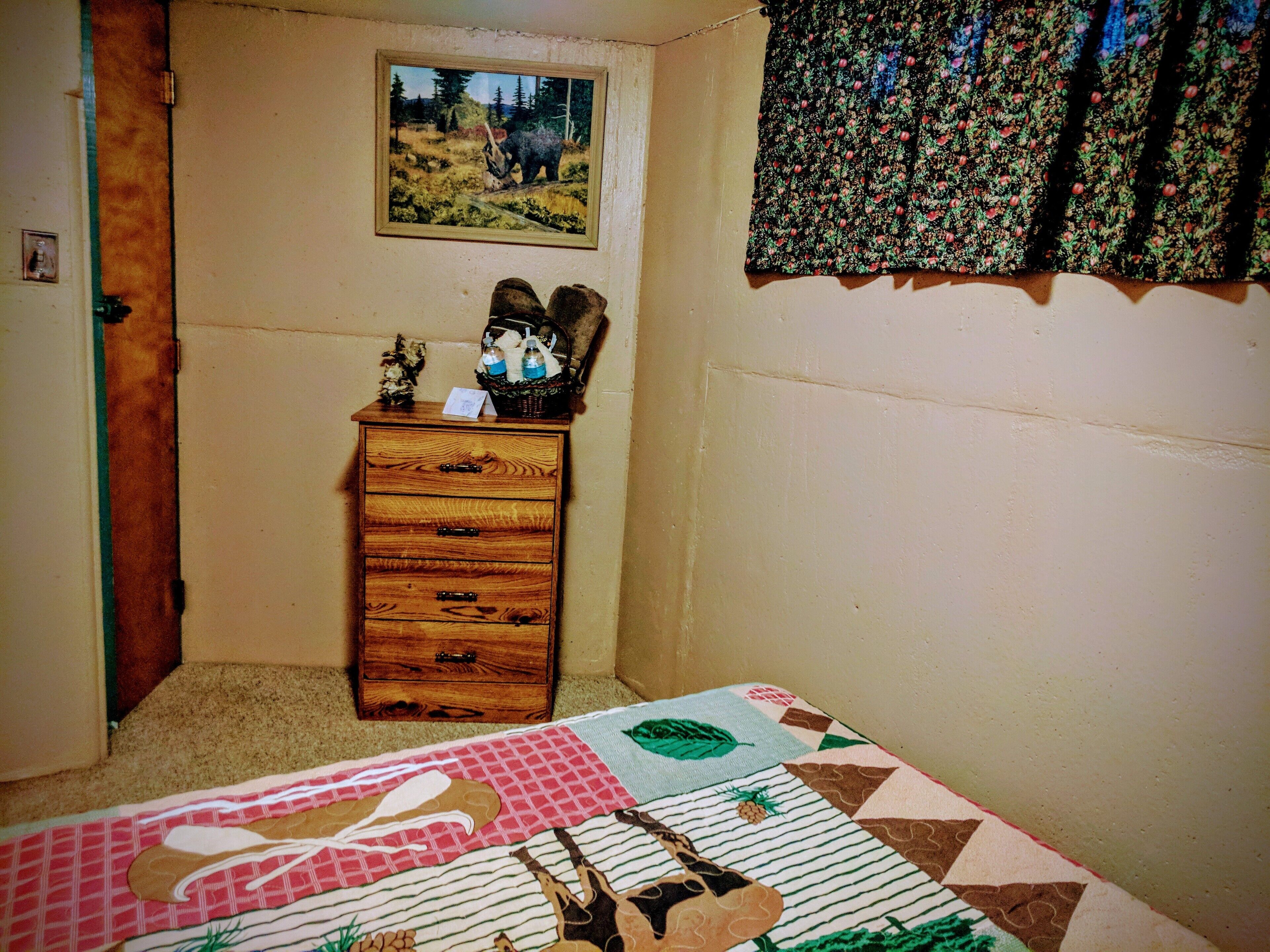 A bedroom with a wooden dresser, a quilt on the bed, a painting on the wall, and a window with curtains.