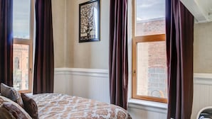 Une chambre à coucher avec un lit, une fenêtre avec des rideaux, un radiateur et un mur de briques visible à travers la fenêtre.