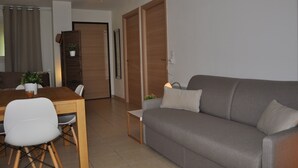 A modern living room with a grey sofa, a wooden dining table, and a white chair. There is a plant on the table and a door in the background. - Charming apartment with large terrace and private pool 5 minutes from the beach (Santa-Lucia-Di-Moriani)