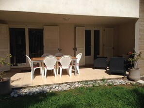 Une terrasse avec une table et des chaises, un pot de fleurs et un petit espace jardin aménagé avec des rochers et de l’herbe.