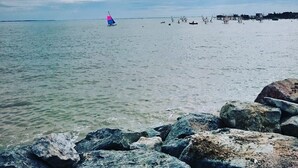Ruhige See mit einem Segelboot in der Ferne, felsige Küstenlinie im Vordergrund und einem Steg mit Booten im Hintergrund.