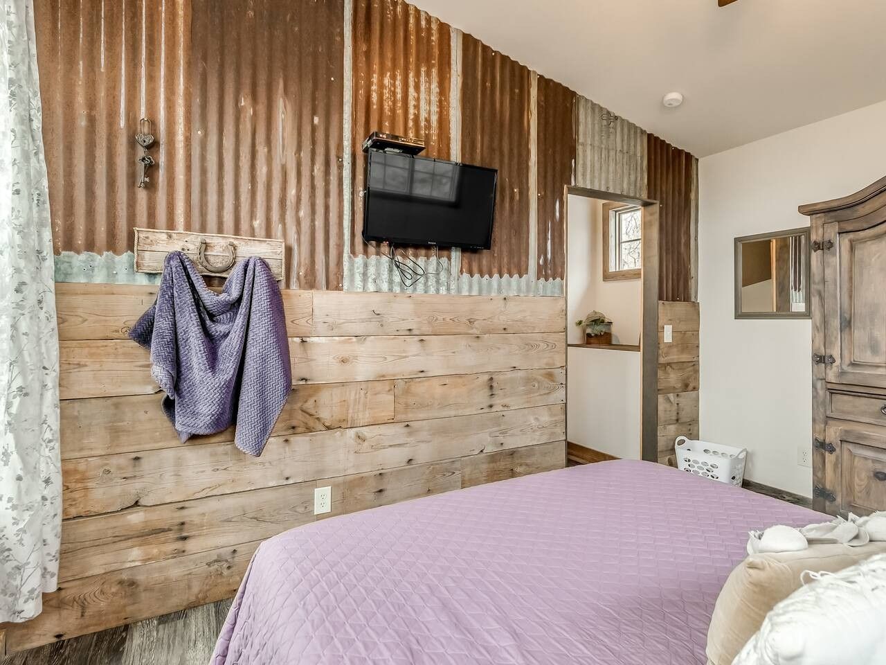 Ein Schlafzimmer mit einem hölzernen Kopfteil, einem Bett mit violetter Bettdecke, einem Fernseher, der über dem Kopfteil angebracht ist, und einem Spiegel an der Wand.