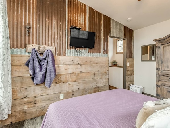 A bedroom with a wooden headboard, a bed with a purple bedspread, a television mounted above the headboard, and a mirror on the wall.