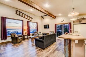 A modern living room with a fireplace, a dining area, and a kitchen with a large window.