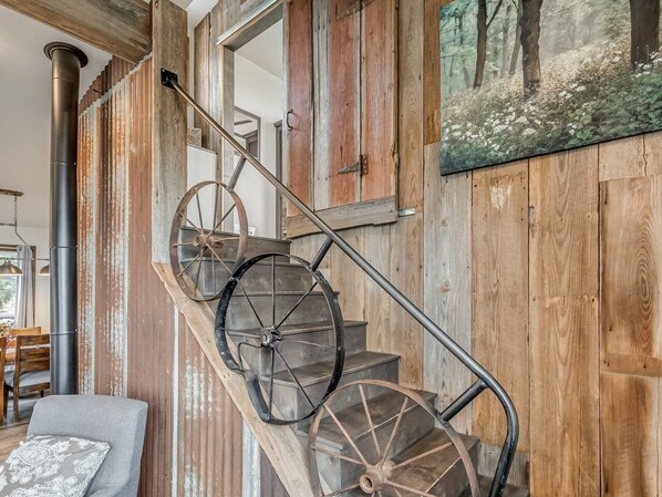 A rustic staircase with a wooden handrail and a large wheel, leading to a room with a wooden wall and a painting of a forest.