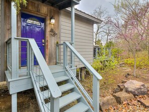 A wooden house with a purple door, a set of stairs leading to a porch, and a garden with rocks and trees.