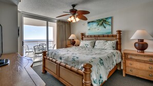 A hotel room with a bed, a desk, a chair, a nightstand, a lamp, a ceiling fan, and a painting of a beach scene. - Sophisticated Beach Chic Unit, Oceanfront Pool | Crescent Shores - S 1106 (North Myrtle Beach)