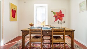 Een eetgedeelte met een houten tafel en stoelen, een kamerbreed tapijt met een patroon, en twee schilderijen aan de muur.