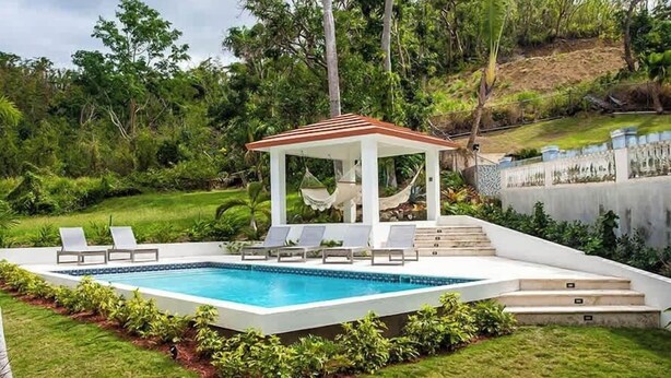 A rectangular swimming pool with a white deck, surrounded by green grass and bushes. There are lounge chairs and a gazebo with a red roof. - R Casa Unit 3 At Steps Beach (Rincon)